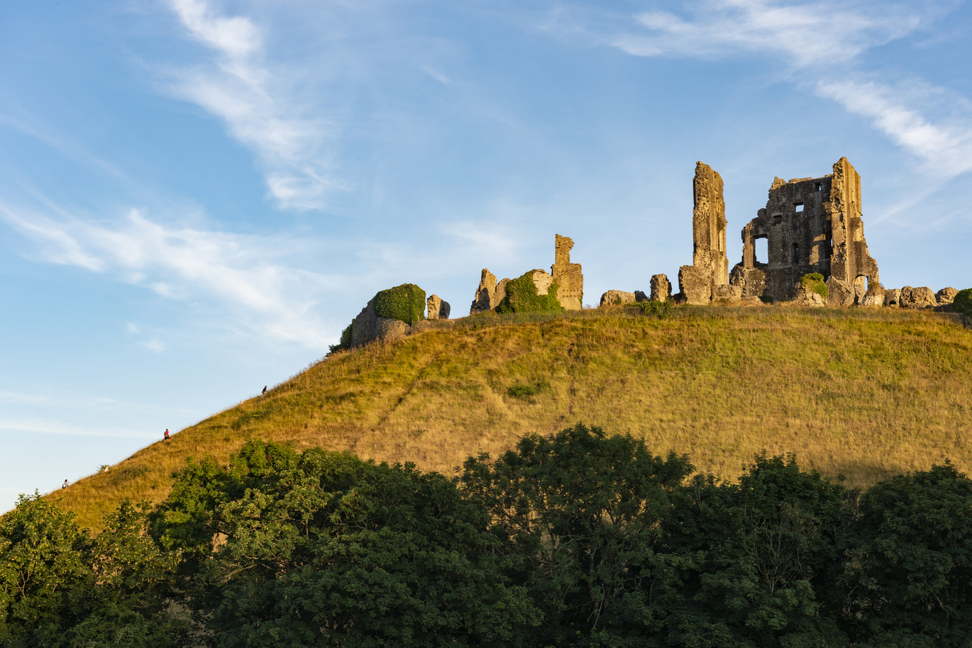 Castle ruins on top of a green hill surrounded by trees and the blue sky with some white clouds above.