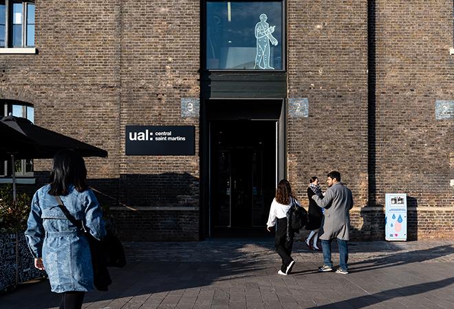 “Entrance to the UAL Central Saint Martins building made of dark brick, with a sign reading ‘ual: central saint martins’ beside the doorway. Several people walk toward the entrance along a sunlit paved area, and a simple white line illustration of a person is visible in a window above the door.”