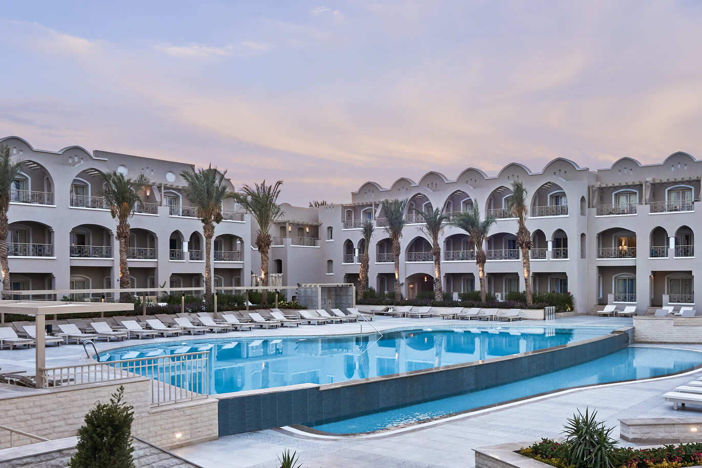 View of the pool area with sun loungers around, surrounded by the white hotel building and the sky with the sun setting above.