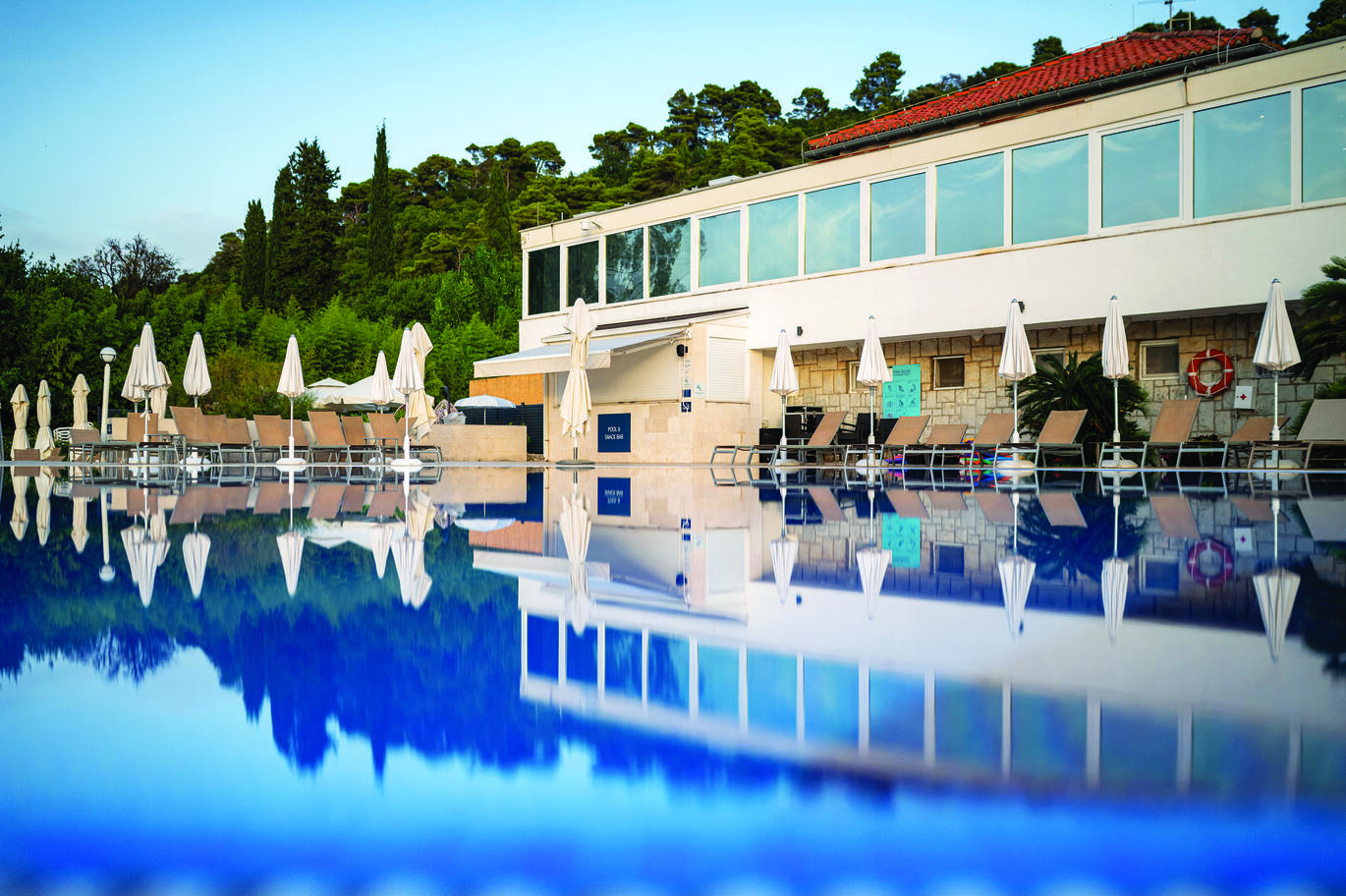 View from the pool of part of the hotel building and sun loungers with umbrellas at the edge. With many trees behind and the blue sky above, all reflecting in the pool.