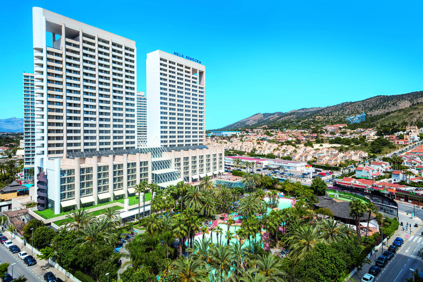 Aerial view of the hotel resort with the tall hotel building at the back and the pools in front surrounded by palm trees. With the resort surrounded by other buildings and mountains in the background on a clear sunny day.