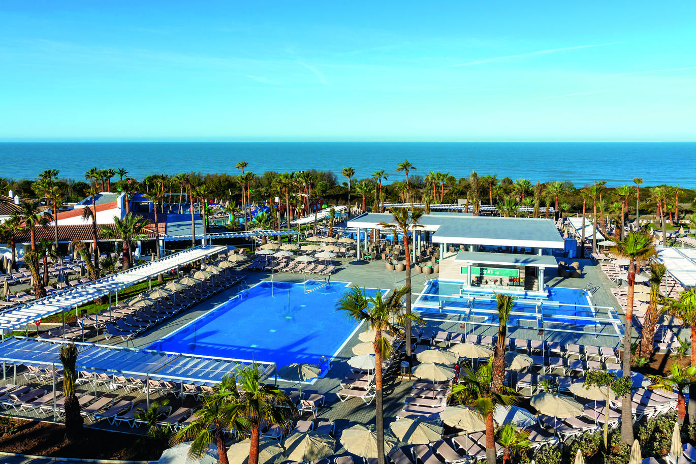 Aerial view of the pool area surrounded by many sun loungers and umbrellas, with lots of palm trees around. With a bar and seating area behind the pools and the sea in the background on a clear, sunny day.