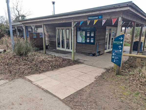 A wooden visitor centre entrance with double doors and a “Welcome” sign is surrounded by paths, plants, and directional signage