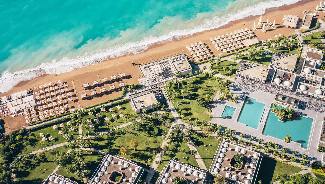 Birdseye view of the pool and lawns scattered with palm trees, with the beach and blue sea with waves crashing onto the beach front.