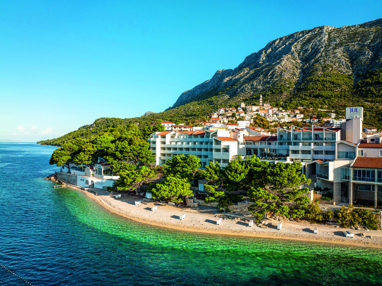 Aerial view over the sea of the beach with trees behind and the hotel building visible behind the trees. With a tall mountain behind covered in trees and the blue sky above.