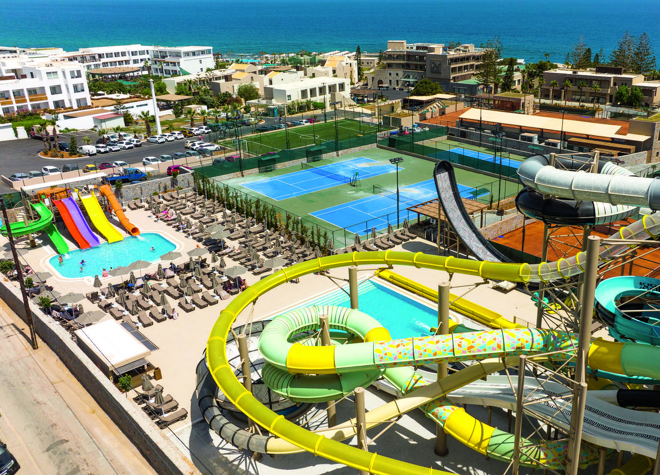View of the waterpark slides with the small pool surrounded by sun loungers. With tennis courts behind and the sea in the background.