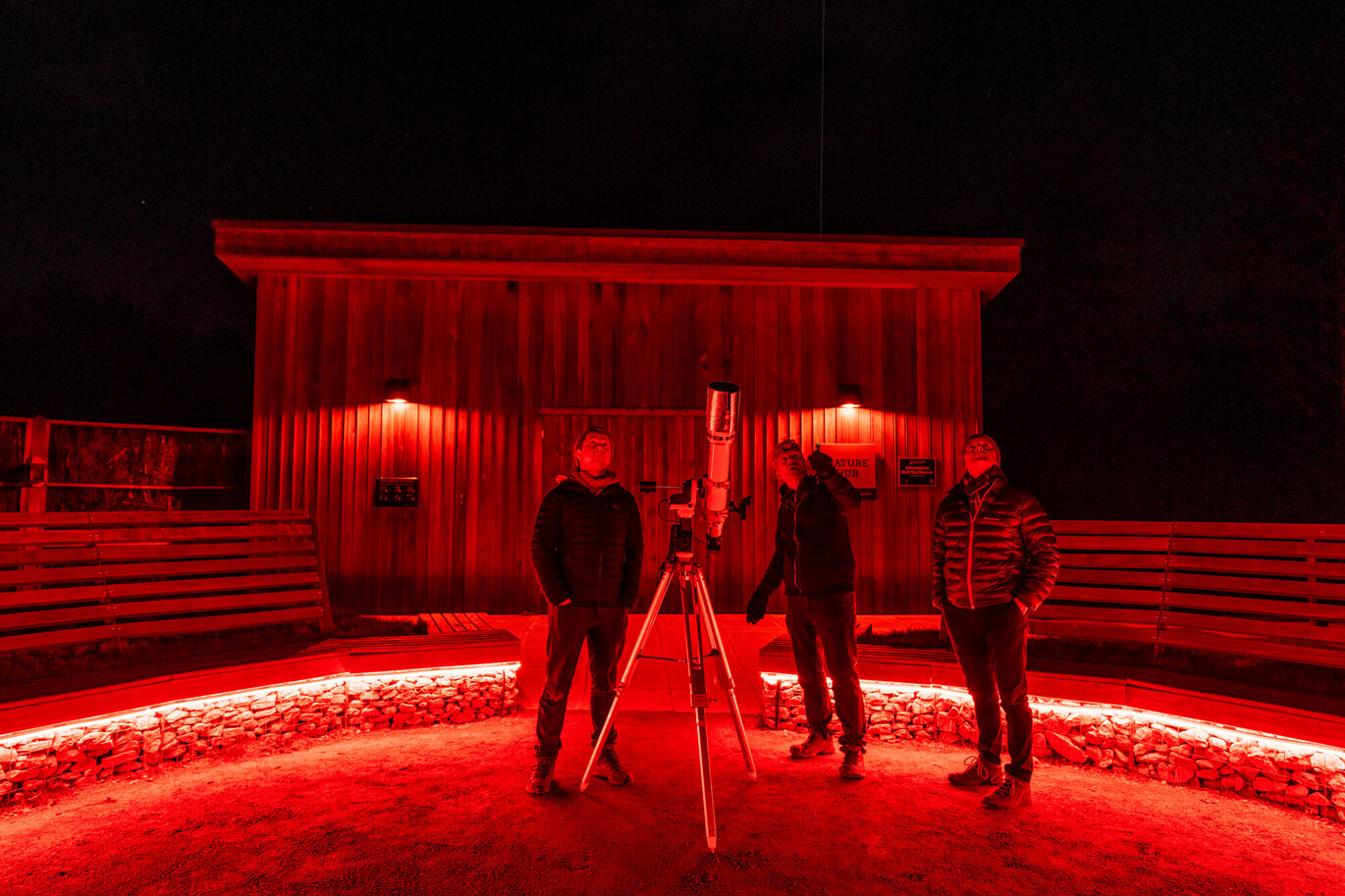 "A wooden building lit by red lights at nighttime with three people stood in front looking at the sky alongside a telescope "
