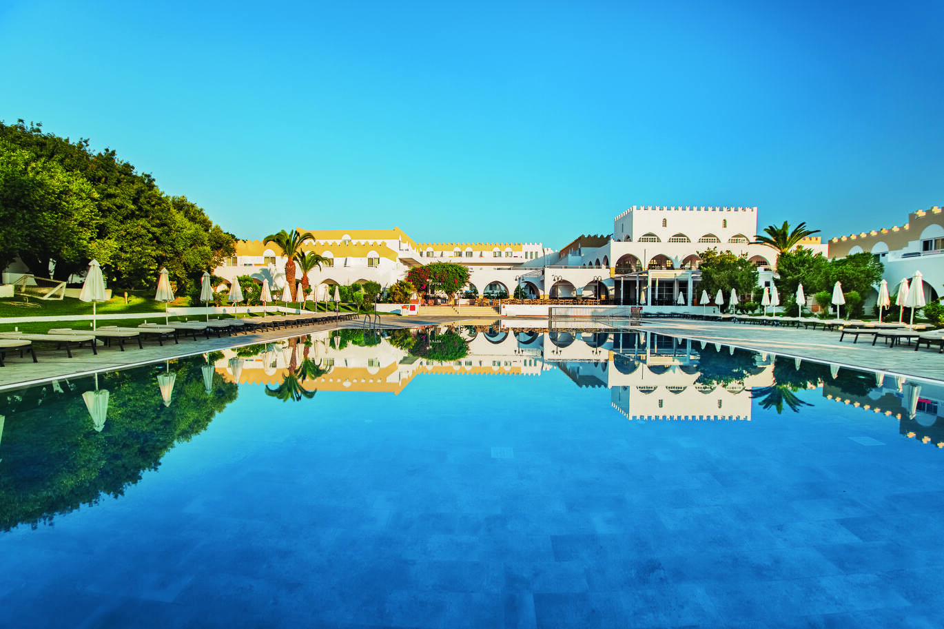 View from the end of the pool of the hotel building and blue sky above reflecting into the pool with trees dotted around and sun loungers around the pool.