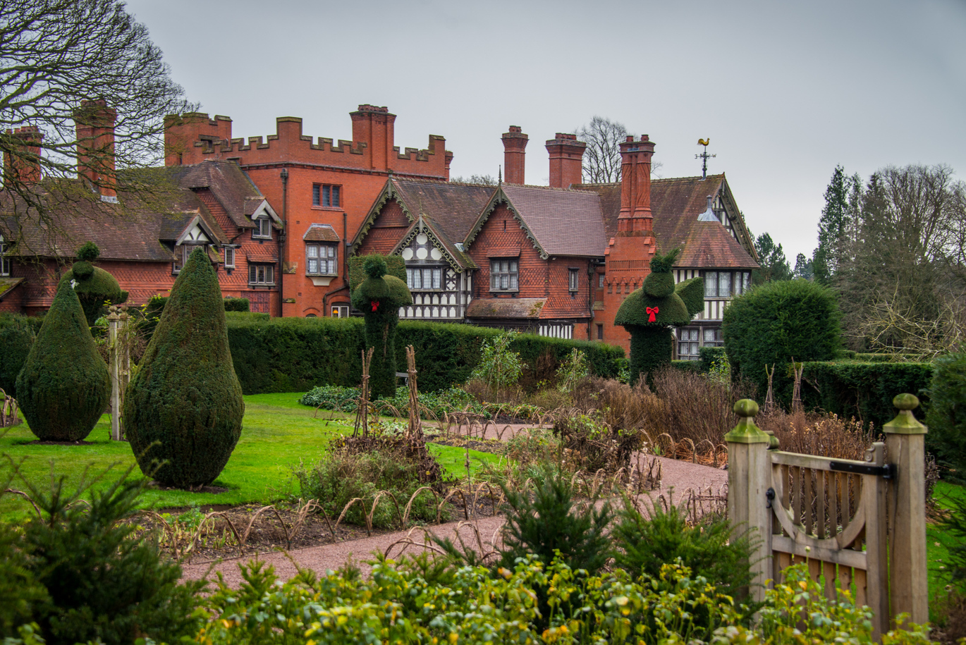 A view of the lush green garden filled with neatly trimmed hedges and a small stone pathway with a small wooden picket gate in the foreground and the large manor house in the background 