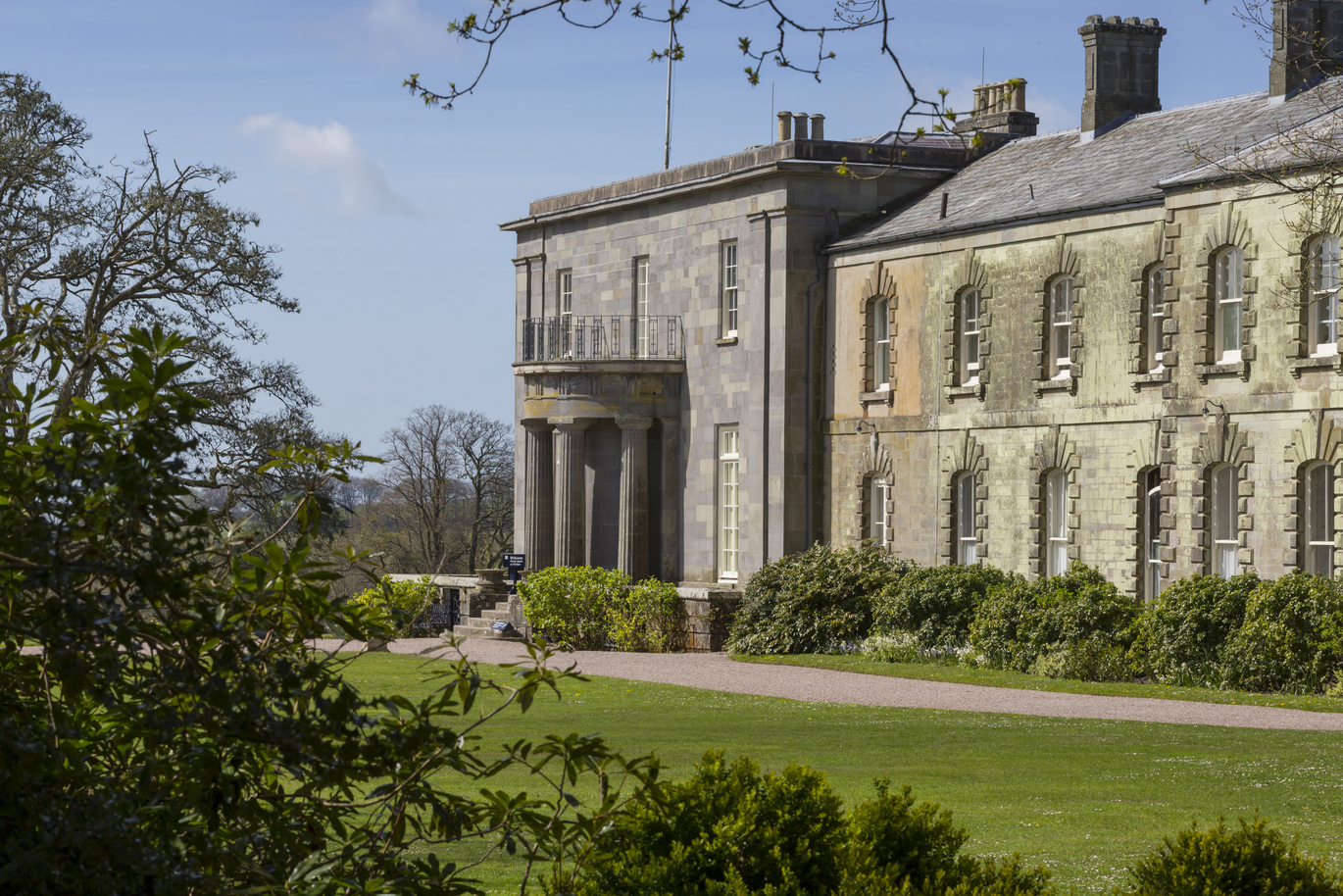 The south front of Arlington Court, Devon. The entrance block of the building is made with grey block stone, the doorway has steps leading up to it and is surrounded by pillars in a U shape with a small balcony at the top. Coming off the right on the entrance building is a long, older style, 2 storey stone building with many arch shaped windows. The front of this building is covered by shrubbery on the ground level along the windows. There is a gravel pathway directly down from the entrance steps which then leads onto a lawn. The picture is taken from behind some trees and you can also see lots of trees in the background. 