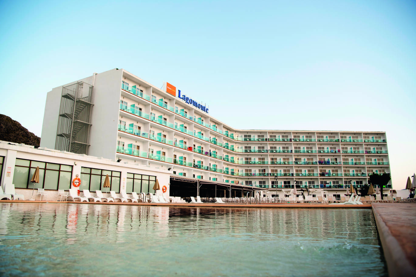 View across the pool of the large building with clusters of hotel rooms with balconies and the name 'Lagomonte' on top of the building. With white sun loungers around the pool and a seating area behind.