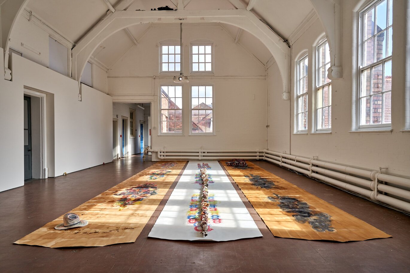 View inside of a hall with a tall ceiling and three long mats laid out on the floor next to each other with different patterns on and large windows around the room.