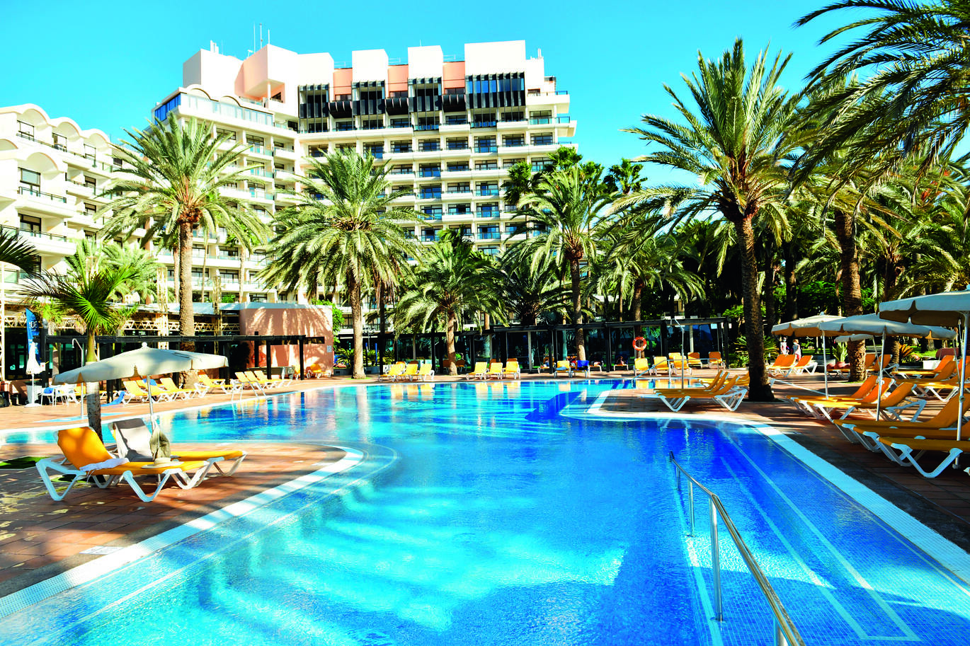View of the hotel pool with the hotel building behind with sun beds and palm trees around the pool.