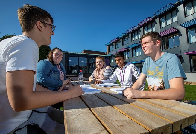 "Photo of 5 people sitting on a bench talking with college building in background"