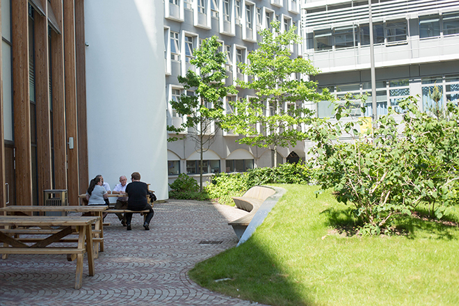 "Outdoor picnic area with students sitting at bench and green area on right hand side and building in background"