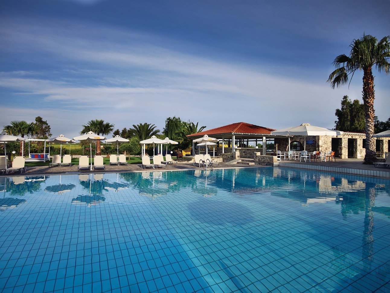 View of the still, empty pool with a few white sun loungers and umbrellas around the edge and some small huts to the side. With some palm trees in view on a sunny day.