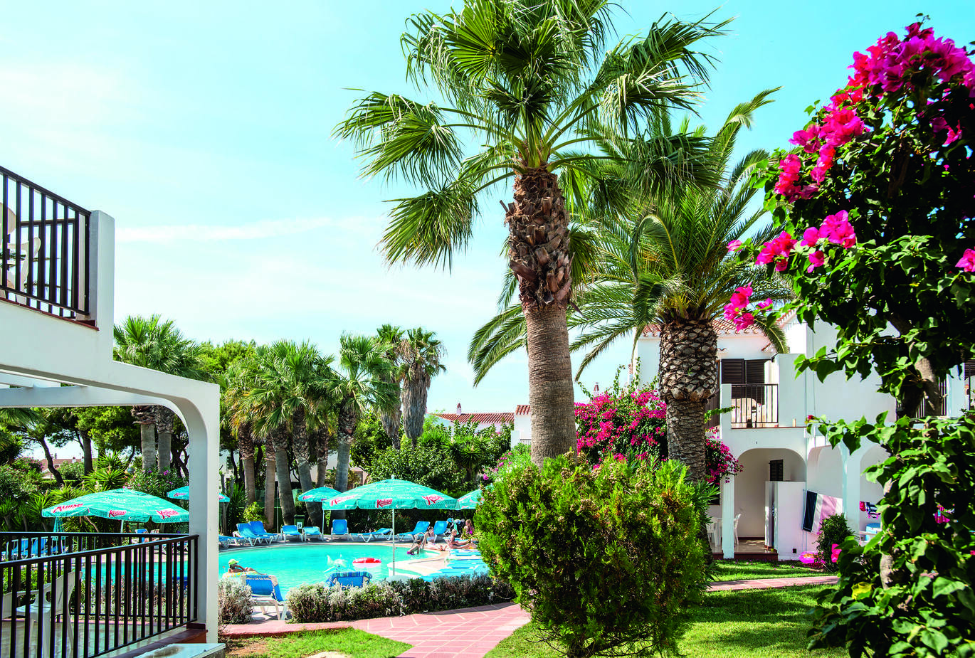 View from behind the pool area between some apartments with palm trees and blossoming trees in the foreground and the pool in the background with people sitting around the pool area on blue sun loungers with blue umbrellas.