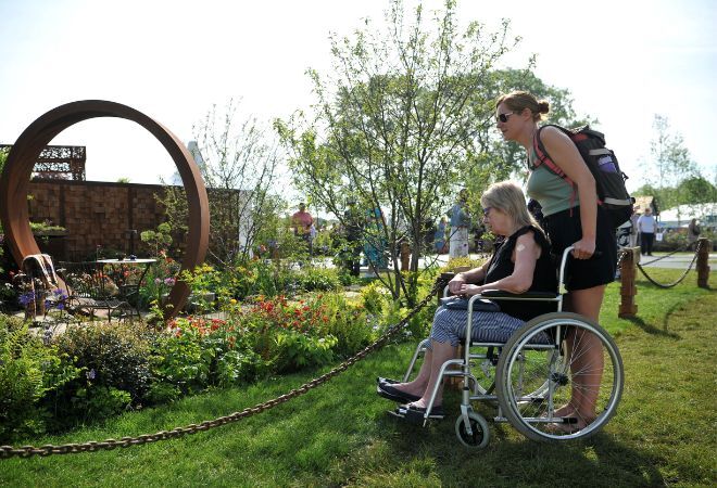 "A femme presenting person using a wheelchair accompanied by a femme presenting person, looking at a garden installation at a show"