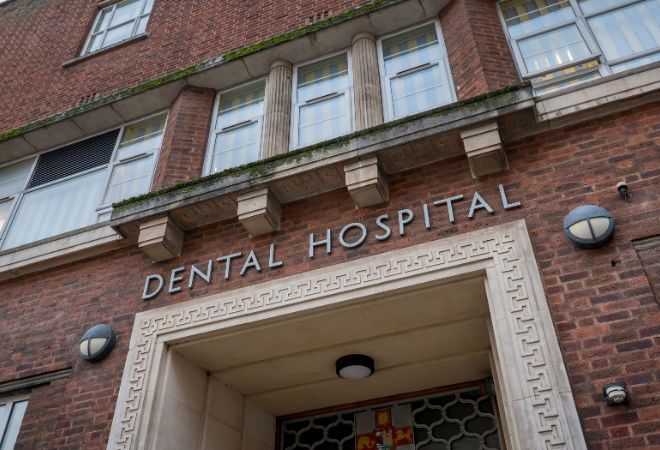 A picture of a sign above the entrance that reads 'DENTAL HOSPITAL' in grey capital letters on the brick background of the building with a wall light either side of the sign. There is an entrance way below the sign with a cement pattern boarder and a light on the ceiling inside. Above the sign is a small ledge with a number of windows above.