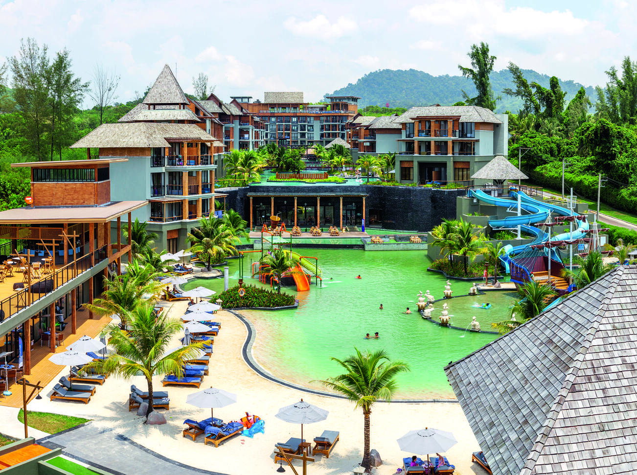 Aerial view of the hotel complex with the pool with waterslides in and sun loungers around. The hotel building is surrounding the pool with trees behind.
