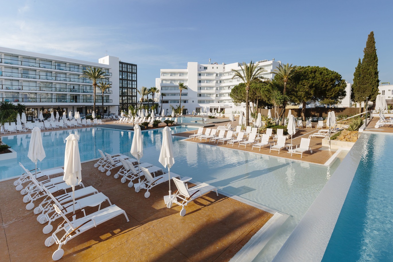 A large swimming pool surrounded with a number of white sun loungers and umbrellas with a few trees surrounding. The white hotel building is behind with the blue sky above.
