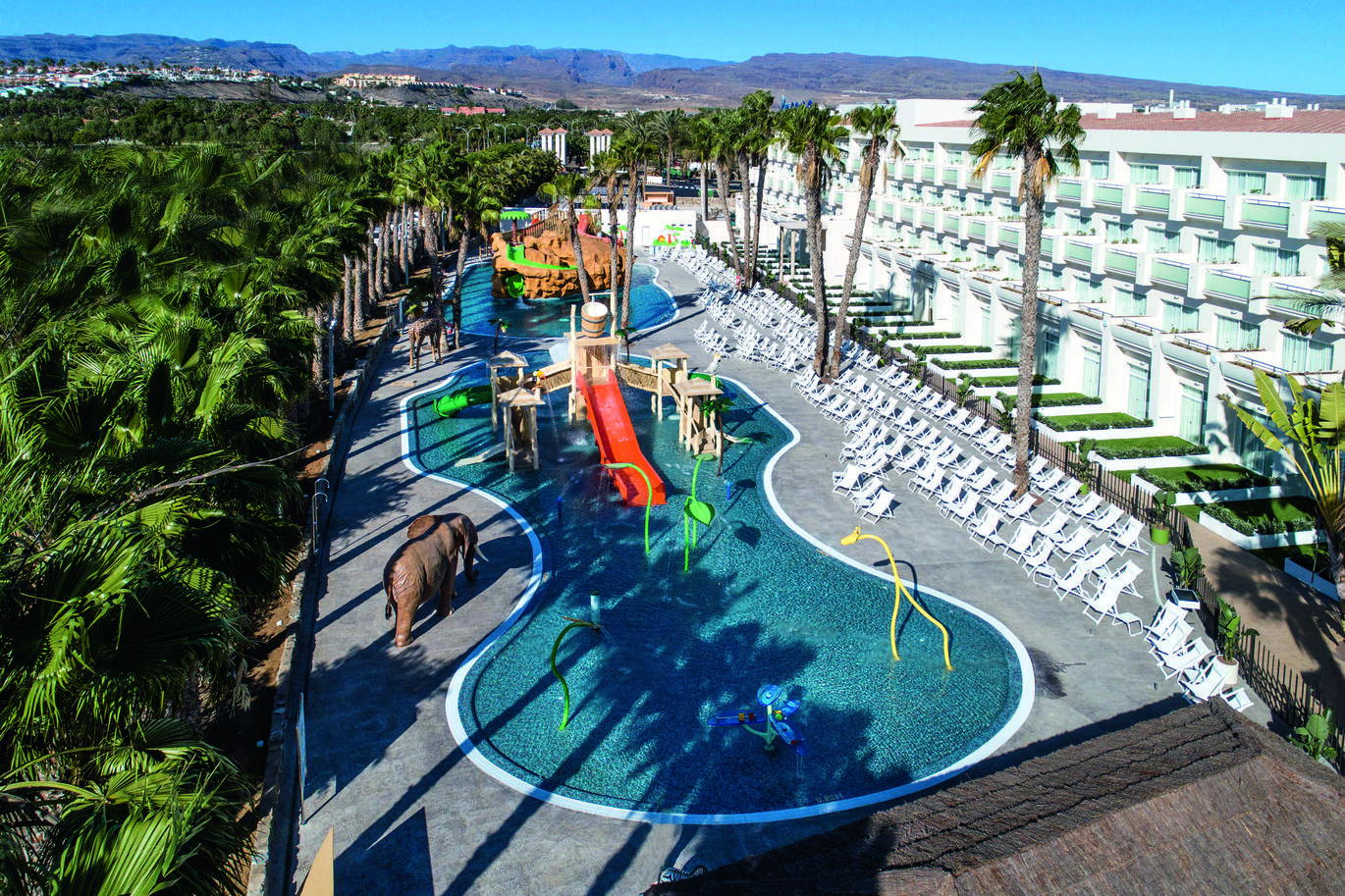 Aerial view of the two swimming pools with slides, climbing frames and play equipment in them. White deck chairs to the right of the pools and to the left is an elephant statue and a forest of palm trees. The white hotel building is to the right.  