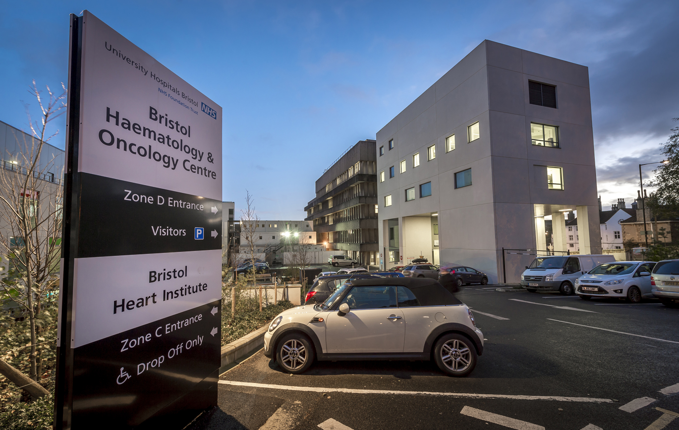 Bristol Haematology & Oncology black and white sign at dusk, within the car park with cars parked in it and hospital buildings behind.