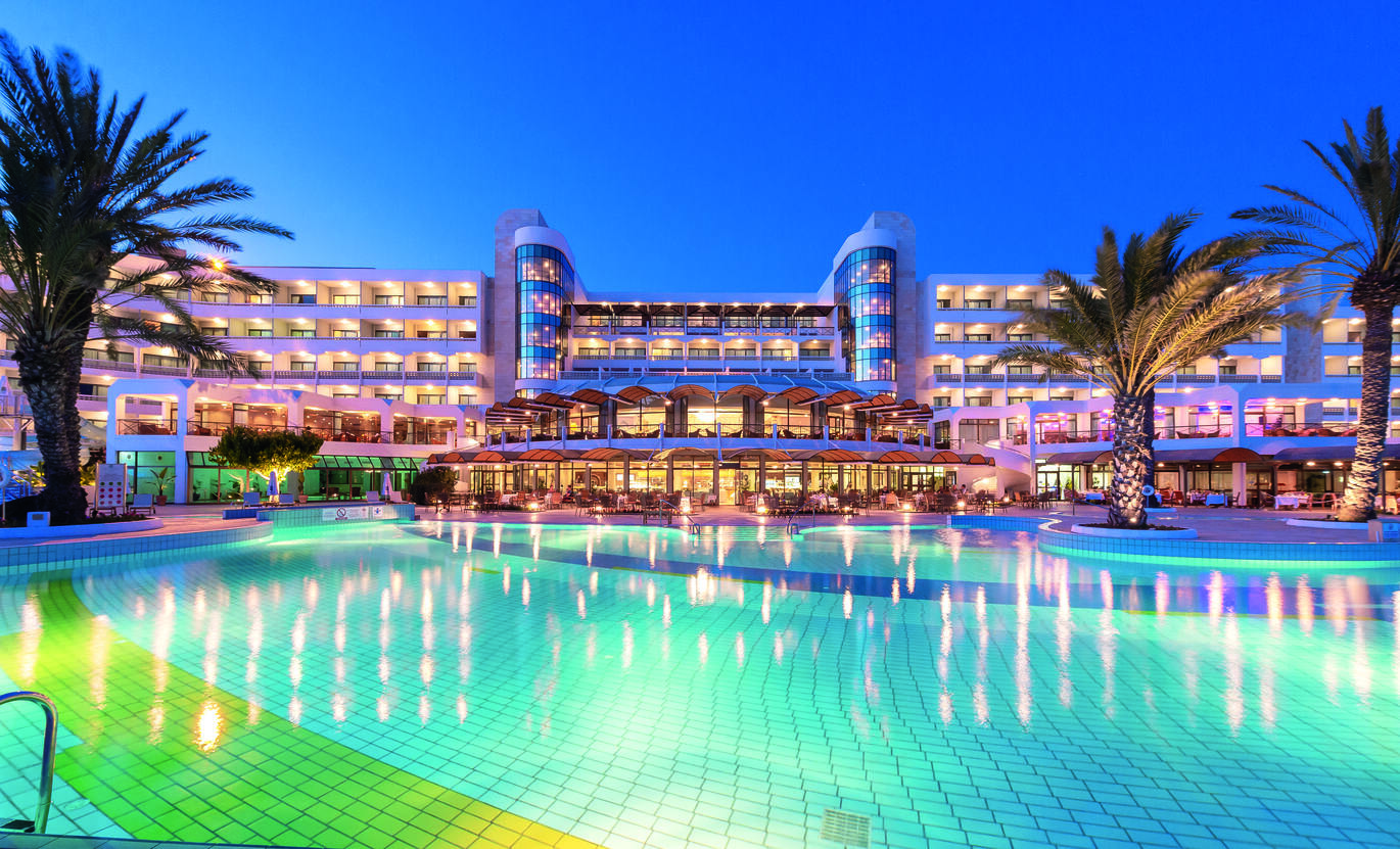View from behind the pool in the dusk of the large hotel building with the lights reflecting in the pool, 