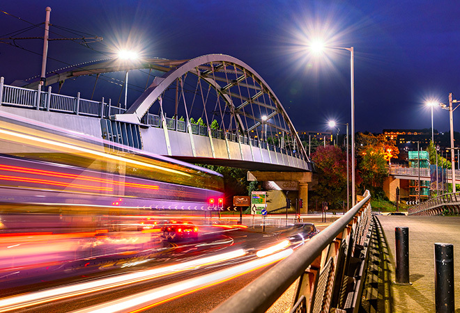 A long exposure shot of cars driving past and under a bridge, nighttime and lots of lights from the cars