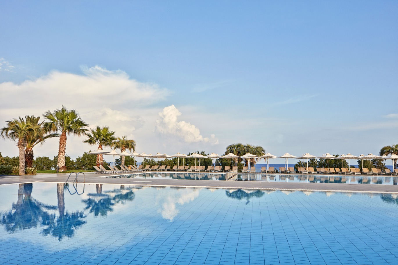 Two large swimming pools surrounded by palm trees reflected in the water and a number of brown sun loungers and white umbrellas. The sea is just visible in the background through the trees. The sky above is blue but slightly cloudy.