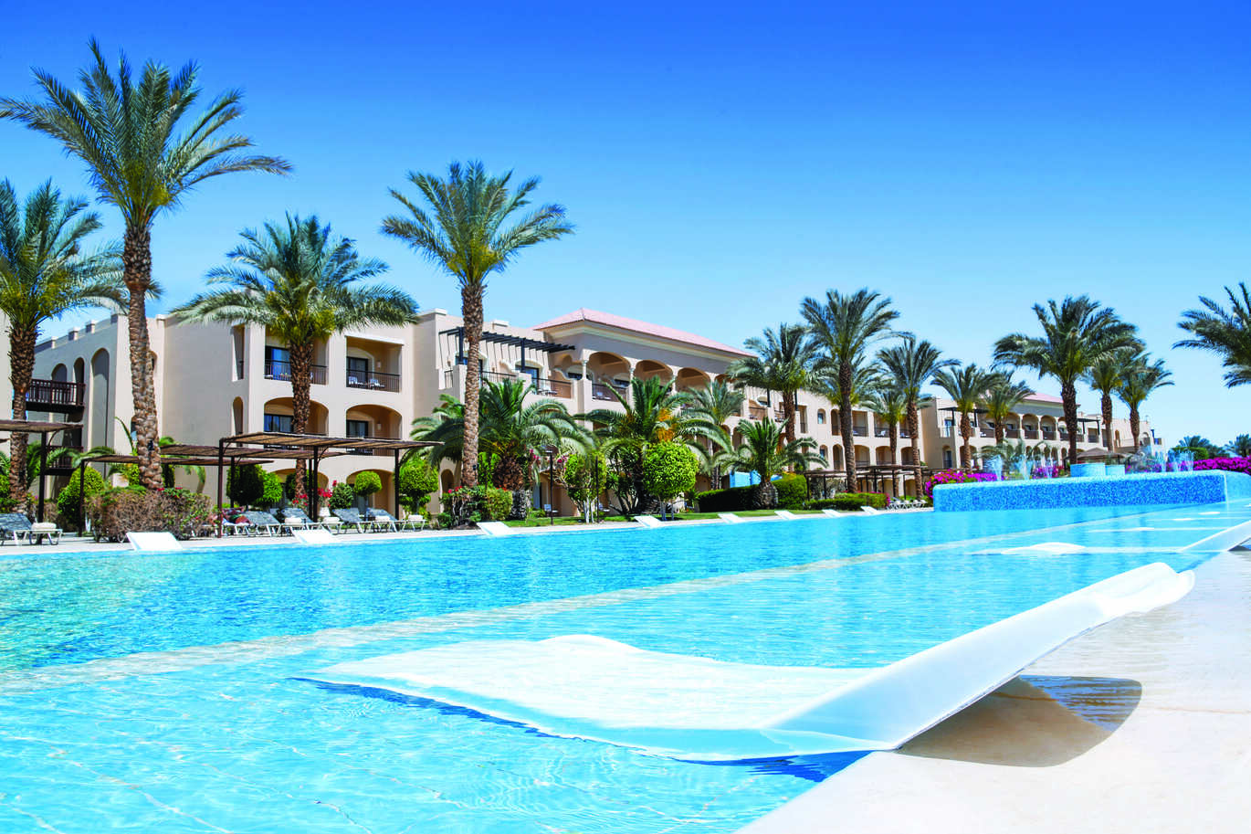 View of the pool area with underwater sun beds lined along each side of the pool with palm trees over the other side and the hotel building behind.