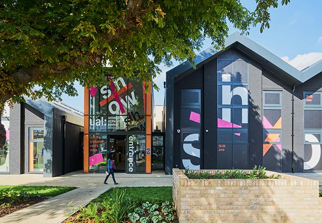 “Modern building with dark gray exterior and large glass entrance framed by orange beams, featuring bold graphic text and colorful geometric shapes on the walls. A tree with green foliage partially shades the foreground, and a brick planter with greenery sits near the walkway.”
