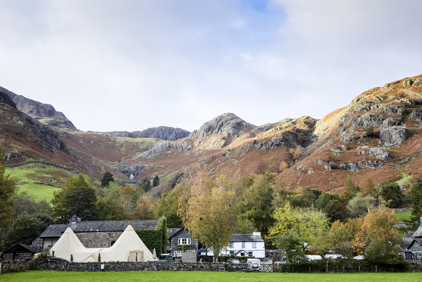 View of a barn and some buildings with a tipi/gazebo type structure with a small stone wall in front leading onto a lush green field. There is a small forest of trees behind which then leads to some magnificent, big hills/mountains. 