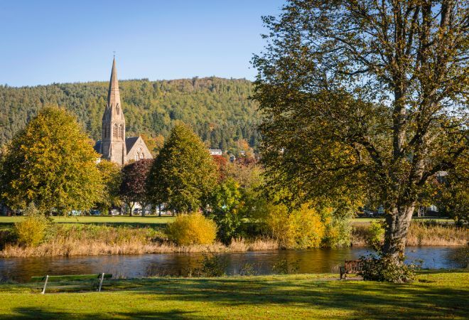 "Peebles on the banks of the river tweed. A river is in the foreground, with grassy banks either side. A church steeple is in the distance, with most of the church hidden by trees"