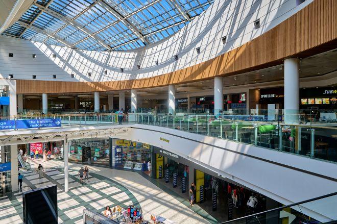 "Bright indoor shopping centre with curved glass roof, upper-level walkway and shops below."