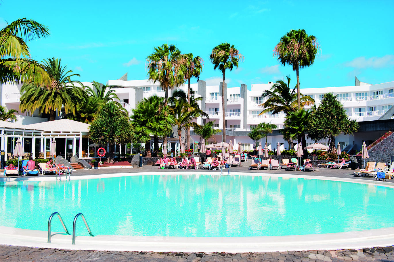 View of the medium sized round pool with people around sitting on sun loungers with palm trees behind and the large white hotel building in the background with the blue sky above.