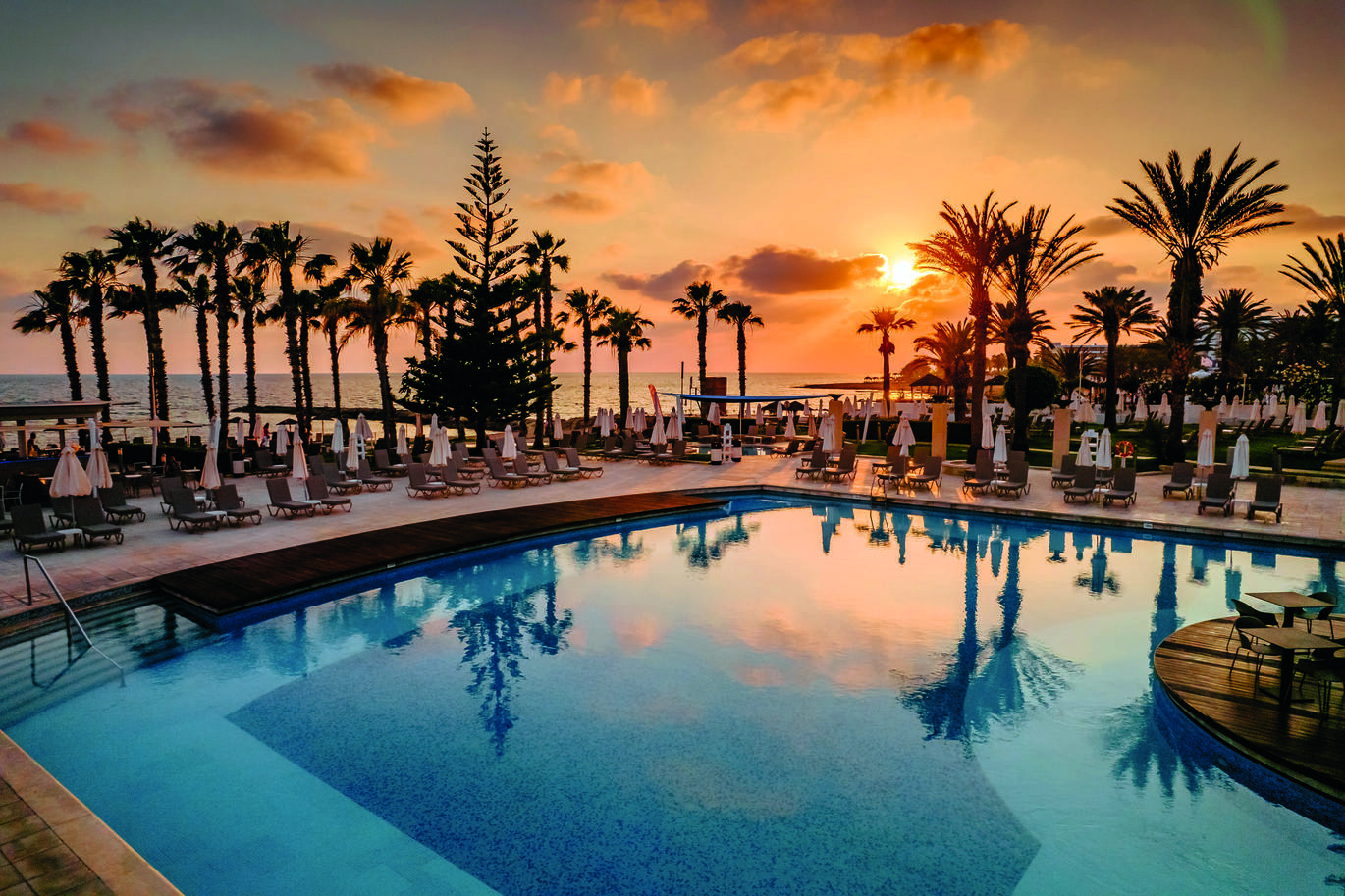 Sunset view of the pool area with sun loungers and umbrellas surrounding, palm trees behind and the sea visible through the trees in the background.