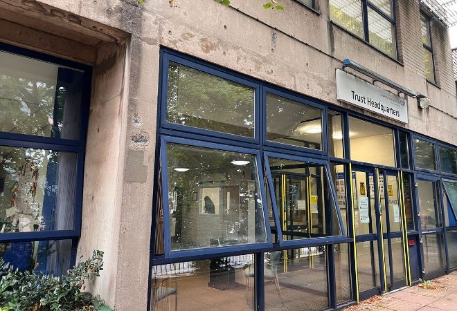 A concrete building with tall windows that have blue boarders and a glass door in the middle of the windows. There is a grey sign above the doors with black writing saying 'Trust Headquarters' with a long light directly above it. 