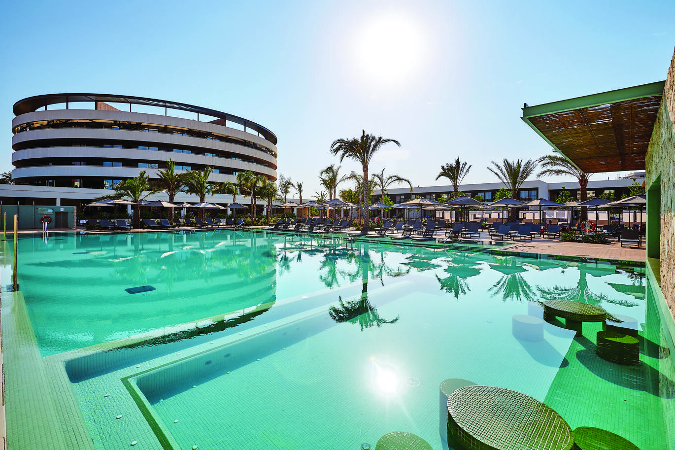 View of the pool area with sun loungers and umbrellas around with tall palm trees reflecting in the pool. The hotel building is behind with the sun shining over the top in the clear, blue sky.
