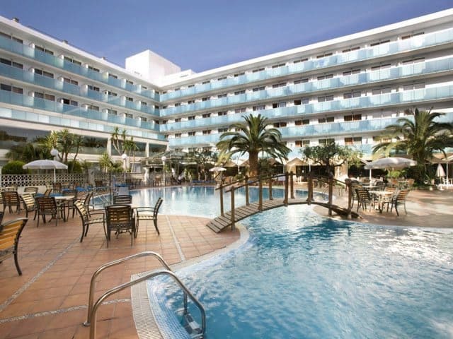 View of the pool area with a small wooden bridge over the middle, with seating areas all around the pool and palm trees dotted around. The hotel building is behind, surrounding the pool area.