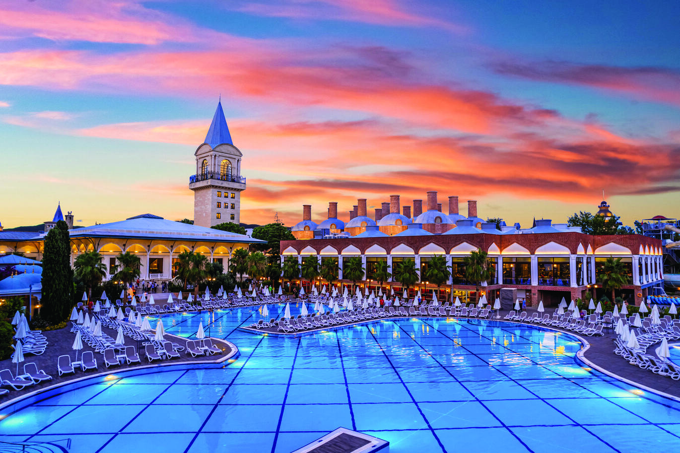 View of the pool area during the sunset, surrounded by many sun loungers with umbrellas and the hotel buildings behind.