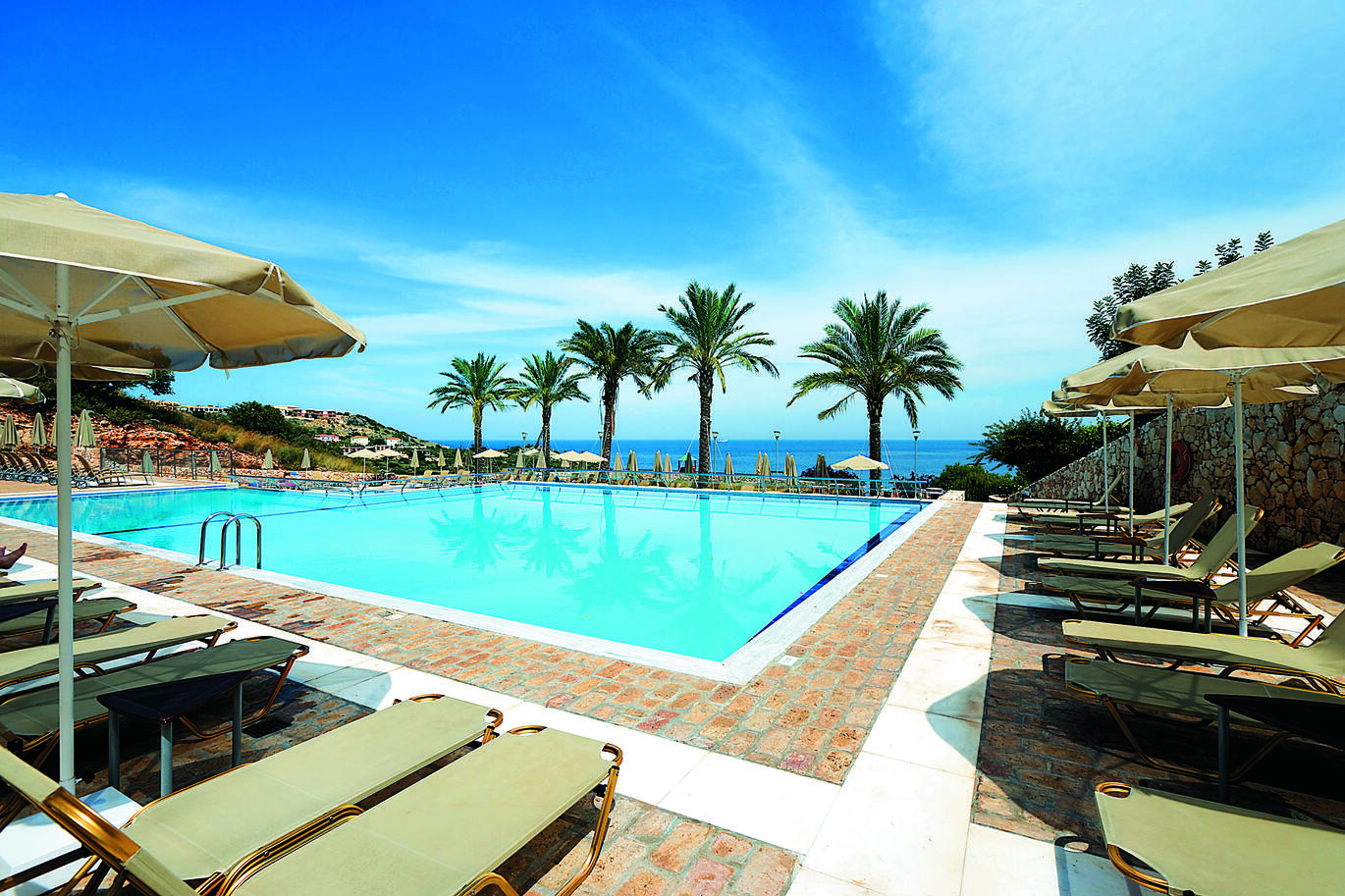 View of the pool area surrounded by sun loungers and umbrellas with a line of palm trees at the back and the sea visible beyond.