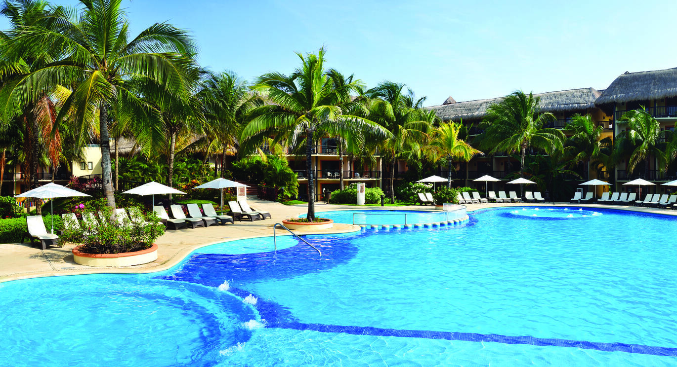 View of the pool with white sun loungers and umbrellas around the pool side with many palm trees dotted around and the hotel buildings just visible through the trees with the blue sky above.