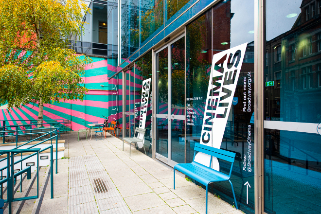 Exterior view of a modern building with a vibrant facade featuring diagonal pink and blue stripes. The glass entrance displays the word “CINEMA” in bold, slanted white letters on black backgrounds. A blue bench sits outside next to a metal railing, and reflections of surrounding trees with autumn-coloured leaves are visible in the glass.
