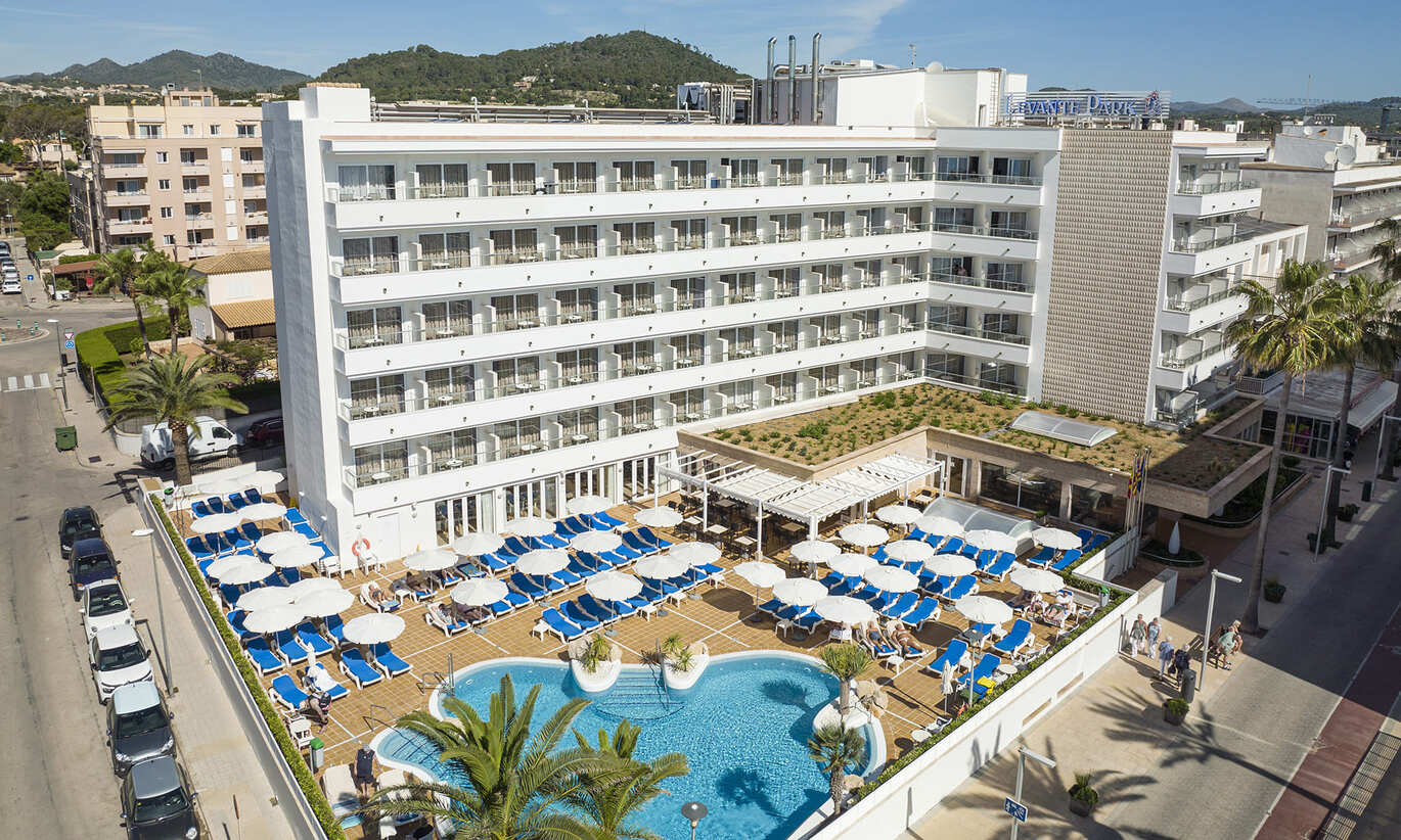 An aerial view of the tall white hotel building with the small blue pool surrounded by blue sun loungers and erect white umbrellas with cars parked on the street outside.