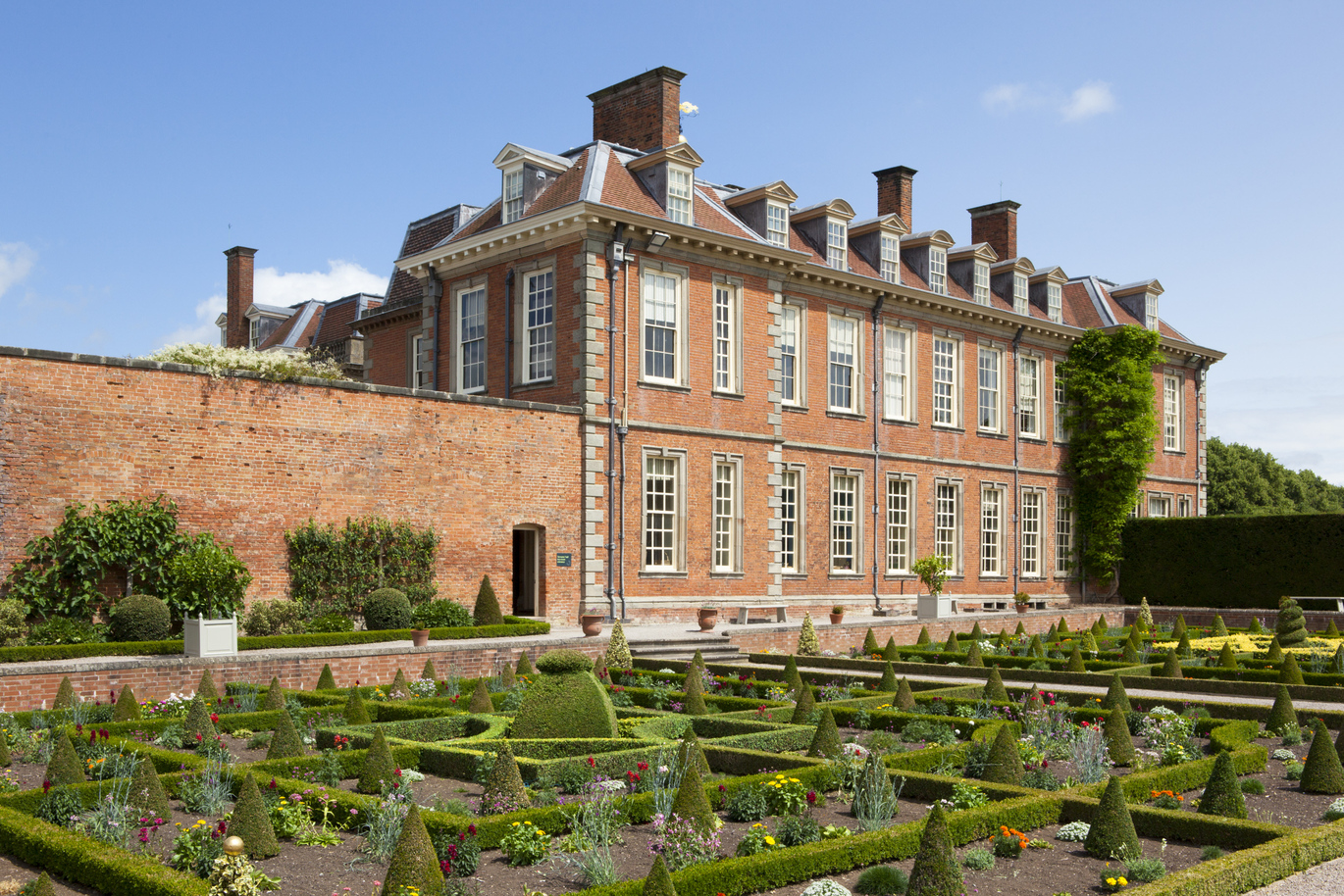 A large brick 18th century house set in formal gardens with a maze of small, neatly trimmed hedges and plants out the front.