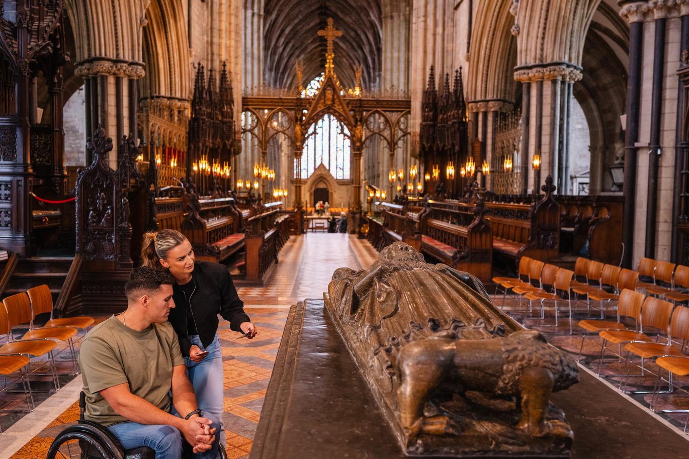 "A man and a woman viewing the resting place of King John buried at Worcester Cathedral. A wheelchair is in use by the man"