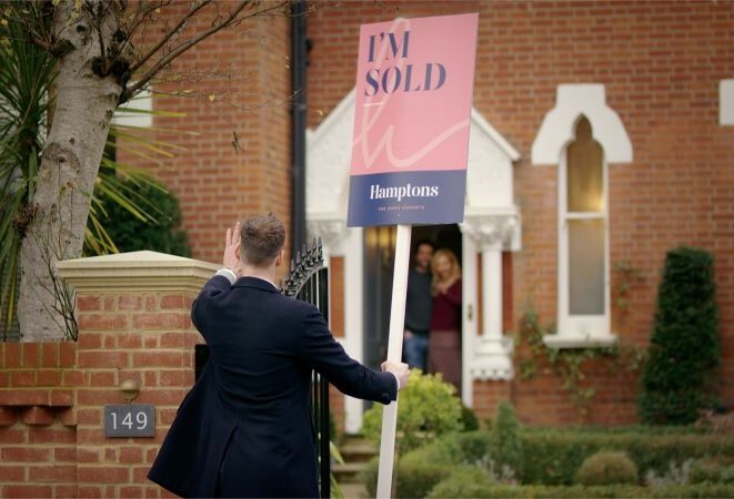 "photo with a Hamptons colleague holding an I'm sold sign, walking up the driveway of a house. A couple are standing in the doorway looking out"