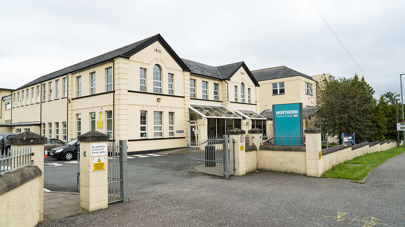 "Exterior image of cream colored building with a pointed roof, cream walls leading in to the building with a teal sign and blue sky in background"