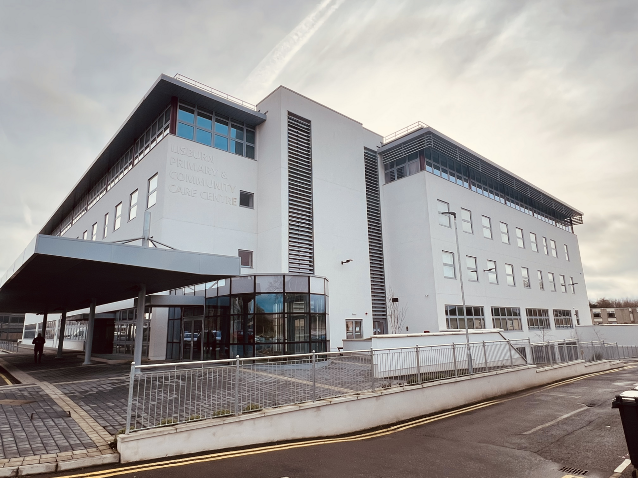 "A large white building with four storeys and signage on the outside reading 'Lisburn Primary and Community Care Centre'. There are metal railings outside and a large canopy over the main entrance and concourse."